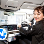 Smiling woman driving a modern car, hands on the steering wheel with a digital dashboard visible ahead.
